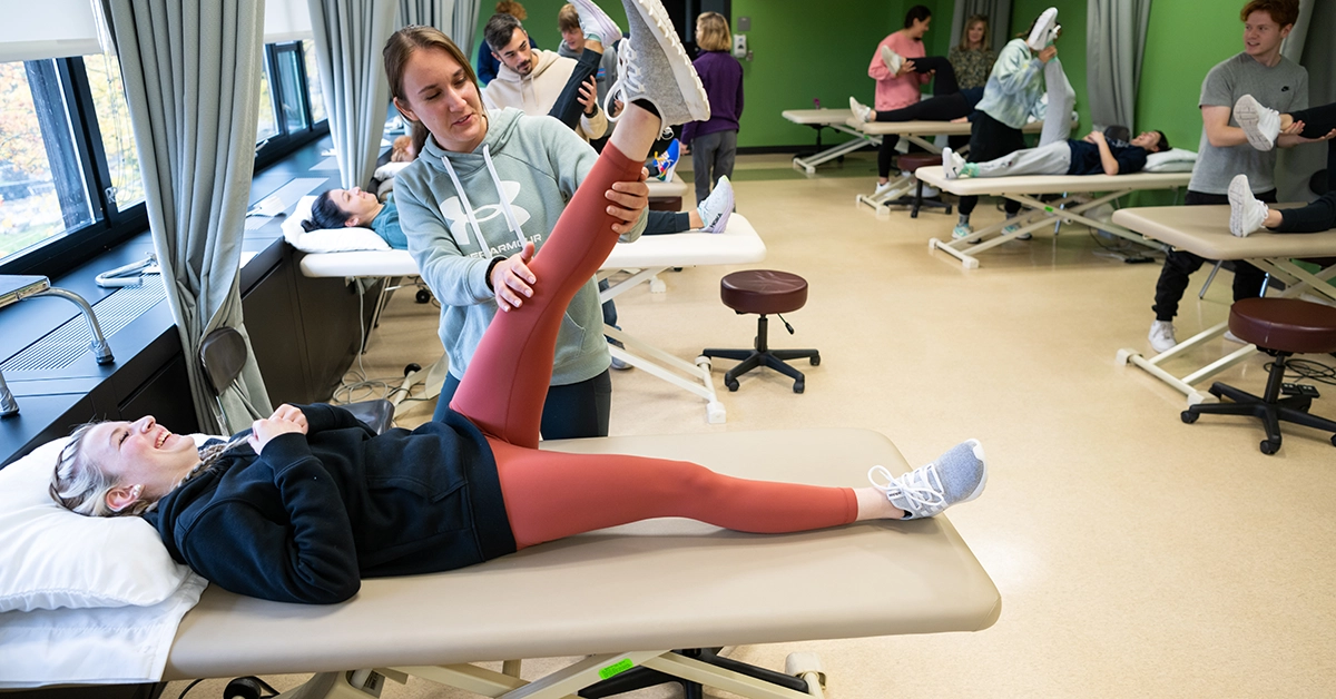 A student helps a student patient stretch their left leg while lying on a table. Other students in the background do the same.