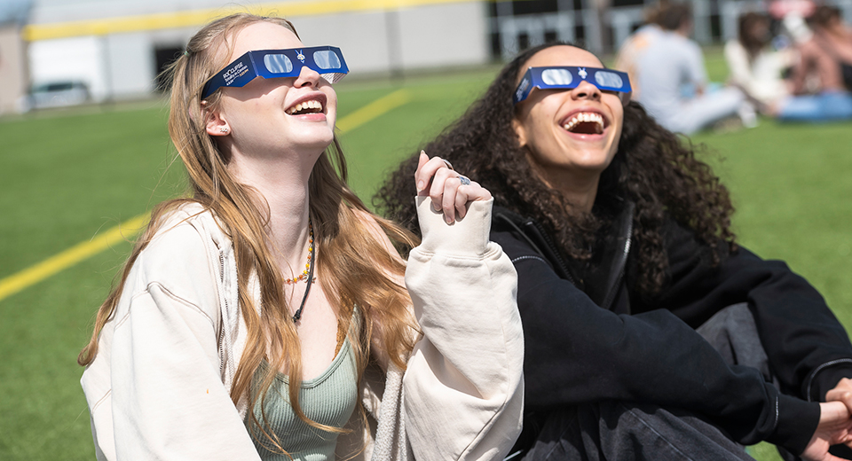 Two seated students wearing eclipse glasses look up at the sun from the turf field.