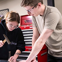 A student holds plywood while another hammers a large nail.