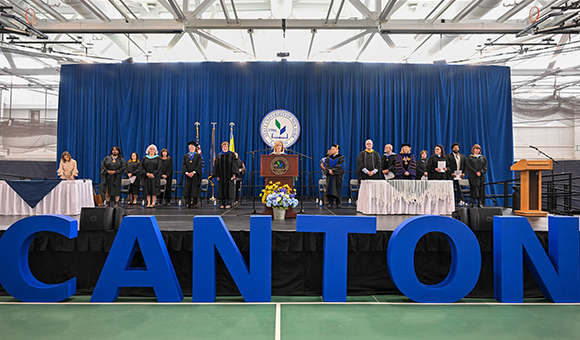 Large blue letters spell Canton in the front of a standing stage party at the start of Honors Convocation 2025.