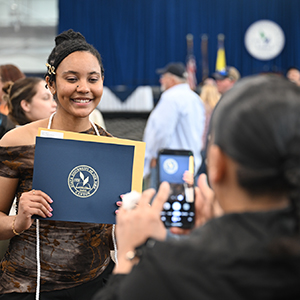 A student proudly poses with an Honors Convocation award while a parent snaps a photo.