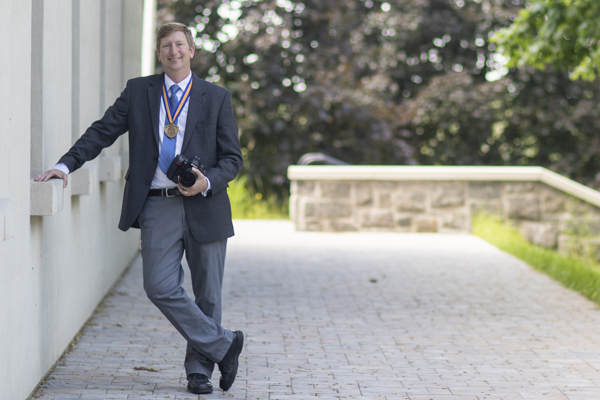 Gregory E. Kie stands wearing his Chancellor's Award medallion and holding a camera outside at SUNY Canton.