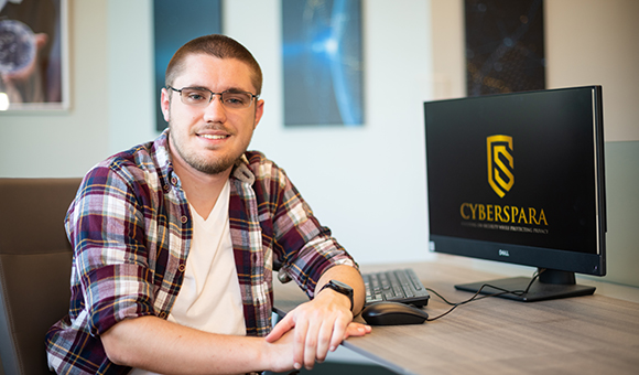 Kyle Meagher sits next to a computer with the CyberSpara logo displayed.