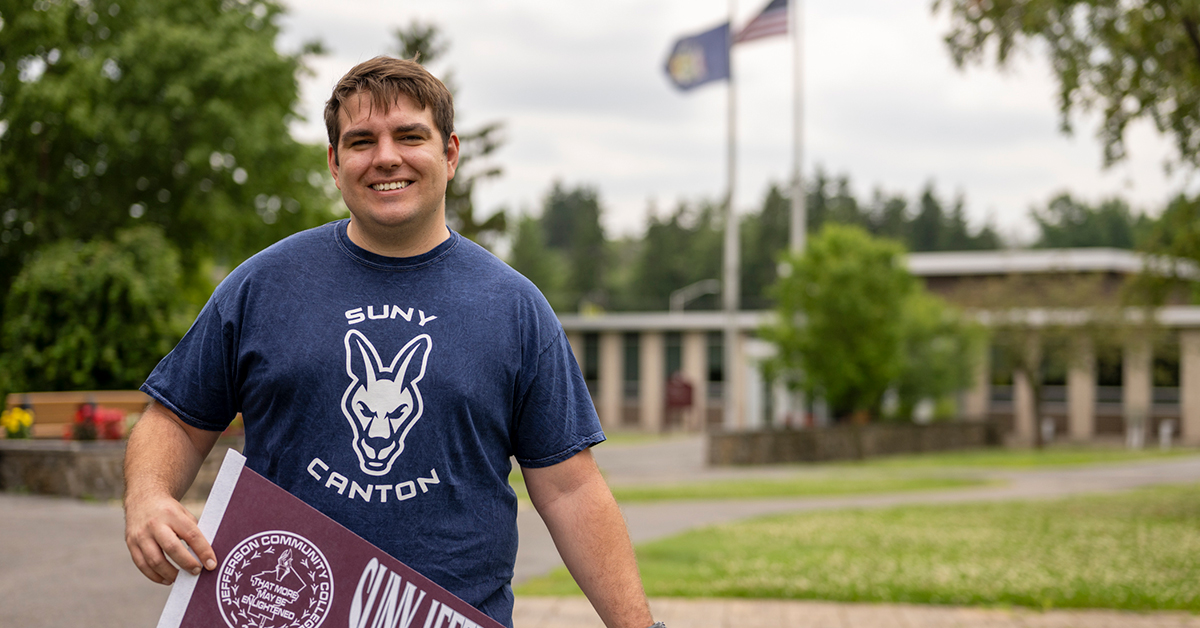 Nathaniel Steelman holds a SUNY Jefferson pennant outside French Hall.
