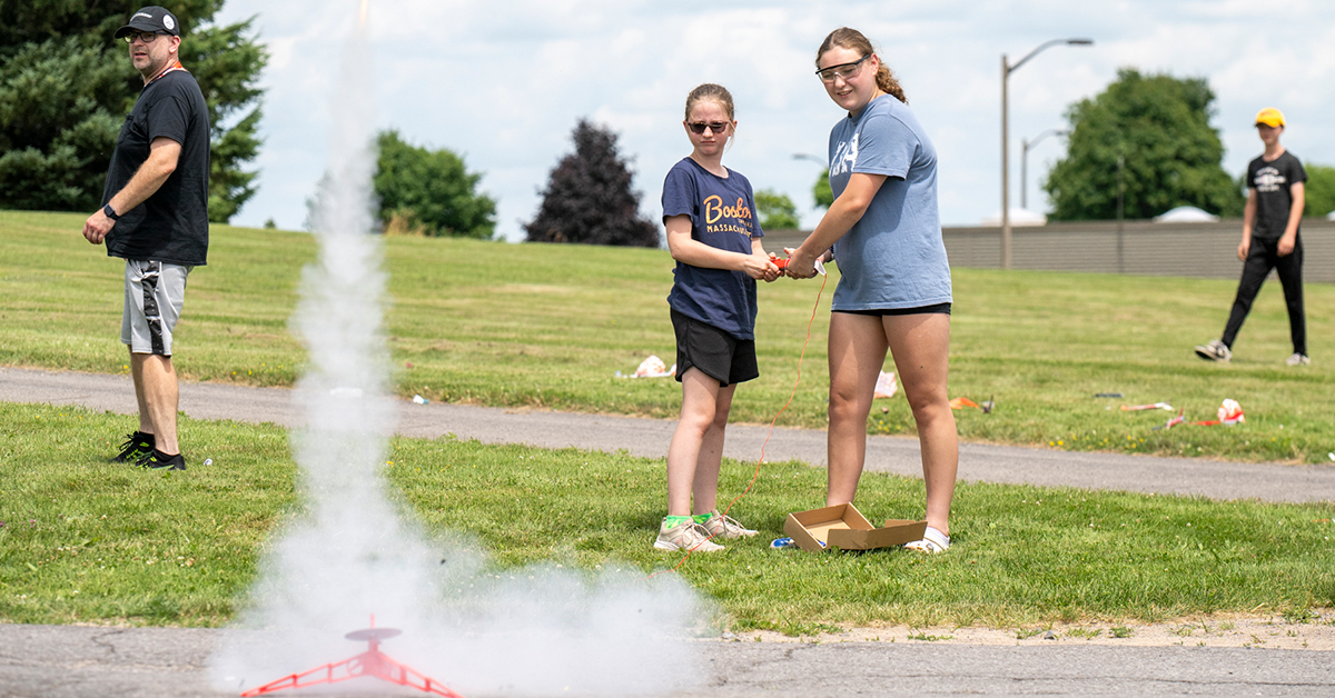Two campers launch a toy rocket outside near the water tower.