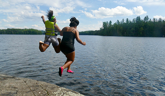 Two students jump into the water with the Adirondacks in the background.