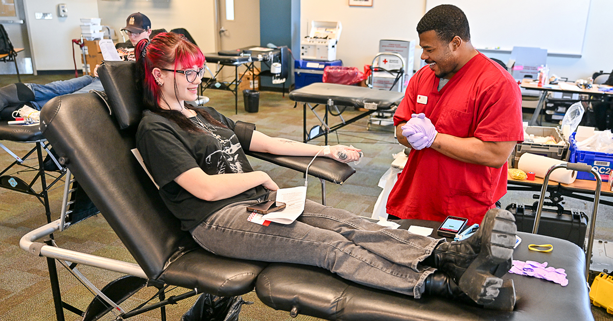 A donor gives blood while a Red Cross member stands nearby.