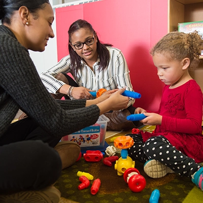 Two child care professionals help a toddler with connection toys.