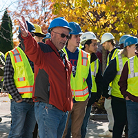 A group of students in yellow vests and blue hard hats get directions from their instructor.