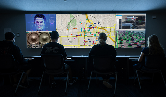 Students work in the crime analysis lab. A large screen depicts a city map while smaller screens depict facial, ballistics, and license plate recognition.