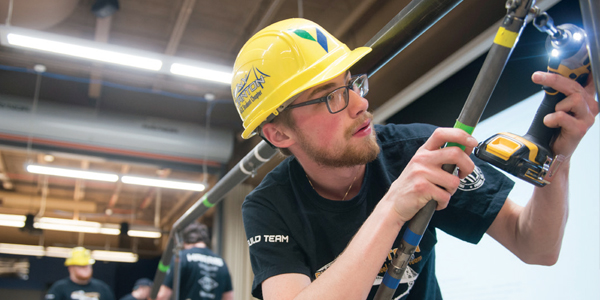 A student with a yellow hardhat drills a bolt into a steel bridge.