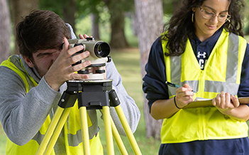 Two students with yellow vests work on surveying equipment outdoors.
