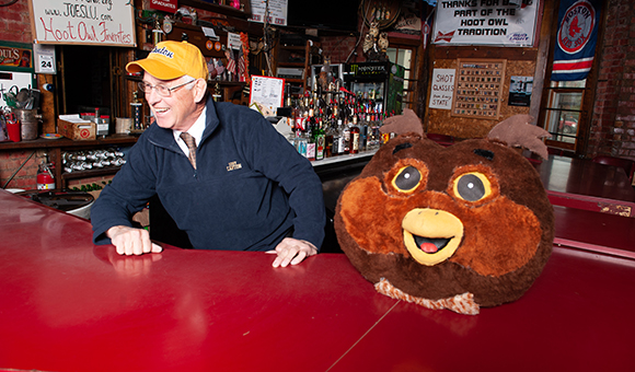 A whimsical stuffed owl head sits on the bar of the Hoot Owl while Danny Fay stands behind sporting his SUNY Canton gear, a yellow hat and blue pullover.