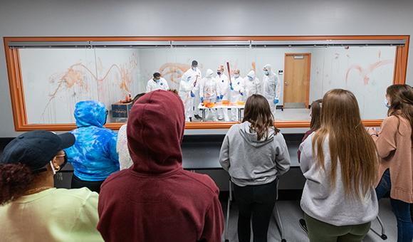 Students participate in a blood splatter demo while classmates watch from the observation space.