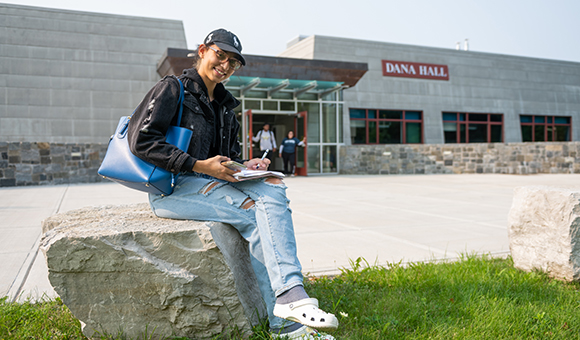 A student takes notes on a rock in front of Dana Hall.