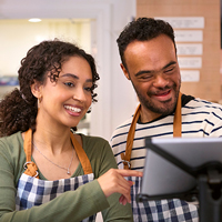 A woman trains a man with down syndrome how to use a checkout system.