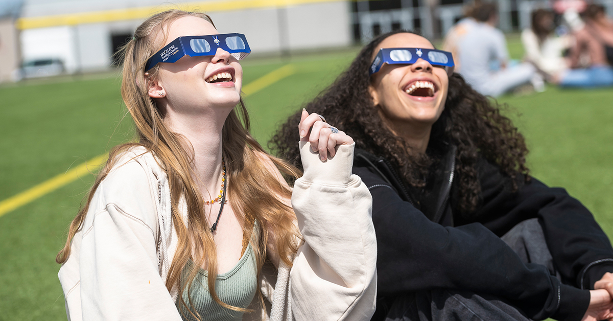 Two seated students wearing eclipse glasses look up at the sun from the turf field.
