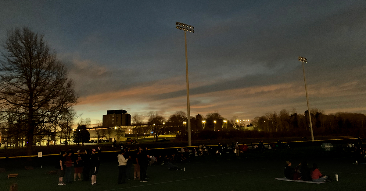 A dusk-like moment during totality. Lights appear around Dana Hall while students watch from the turf field.