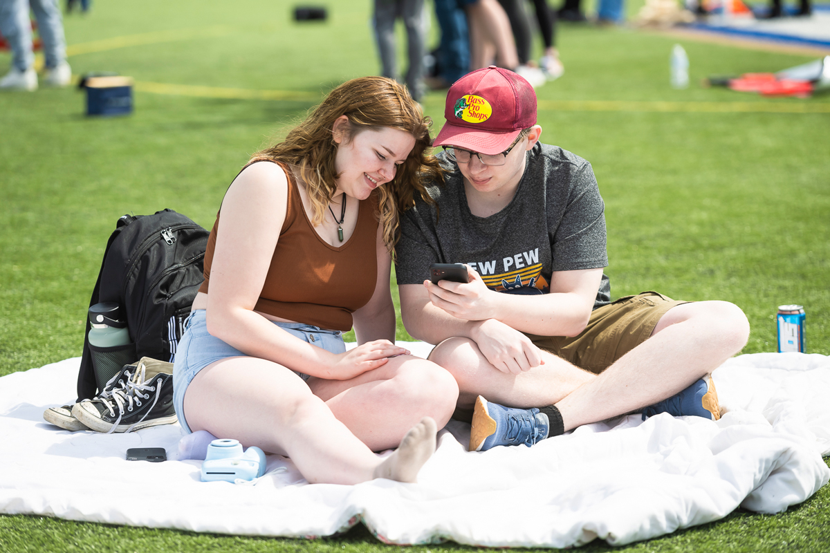On the turf field, two students sit on a blanket while sharing a cell phone.