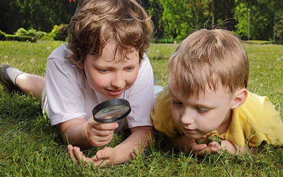 Two campers use a magnifying glass outside to look at insects in the grass.