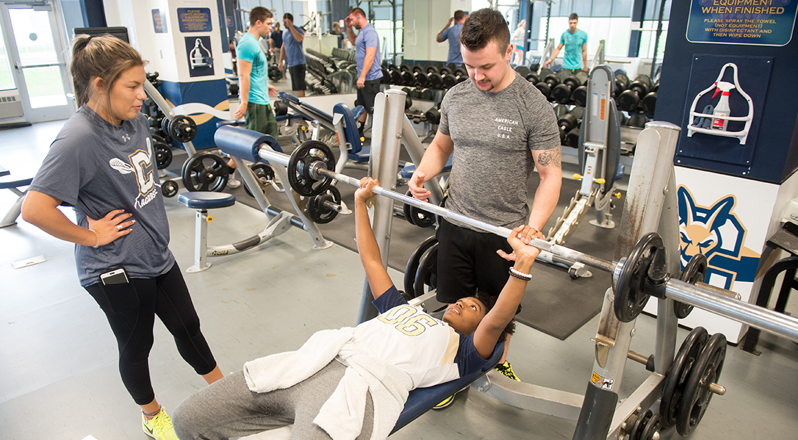A student bench presses in the Fitness Center while another spots from behind and a third watches the form.