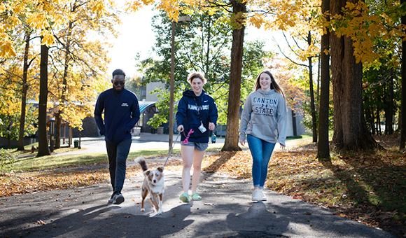 Three students walk a dog from the residence halls on a fall day.