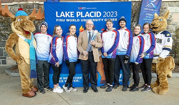 President Zvi Szafran holds the FISU torch while Mac the Moose, Roody Roo, and SUNY Canton athletes flank him.