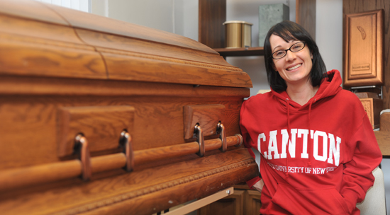 A student in a red SUNY Canton hoodie stands next to a wooden casket.