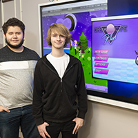 Two students stand next to their developed video game home screen.