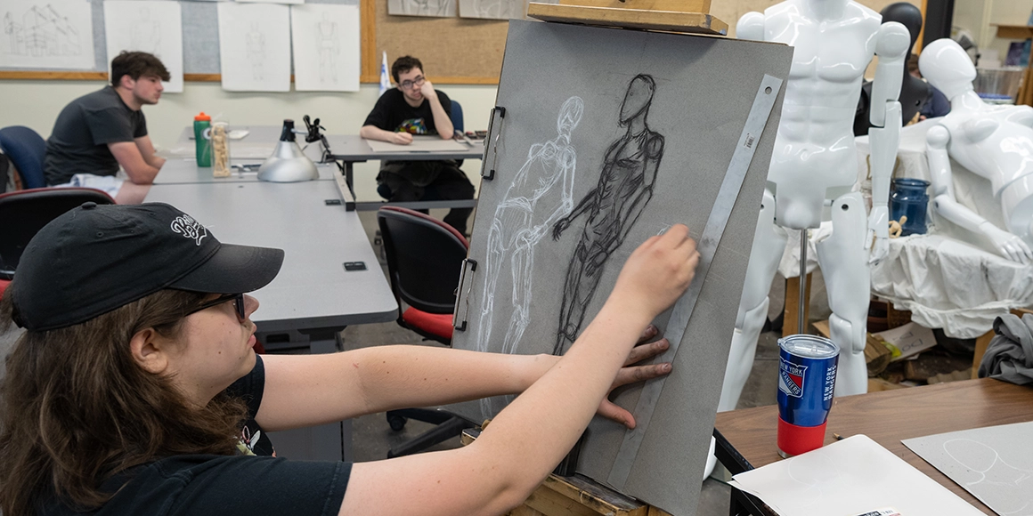 A students uses charcoal to sketch a human figure on an easel.