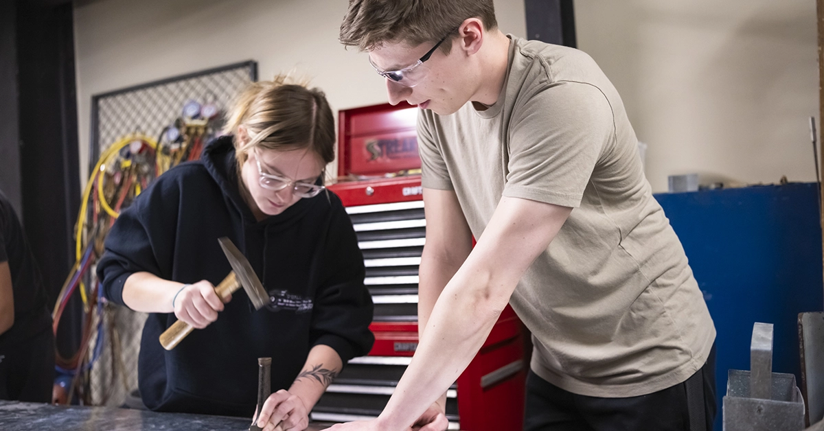 A student holds a large spike while another hammers it into a sheet of metal.