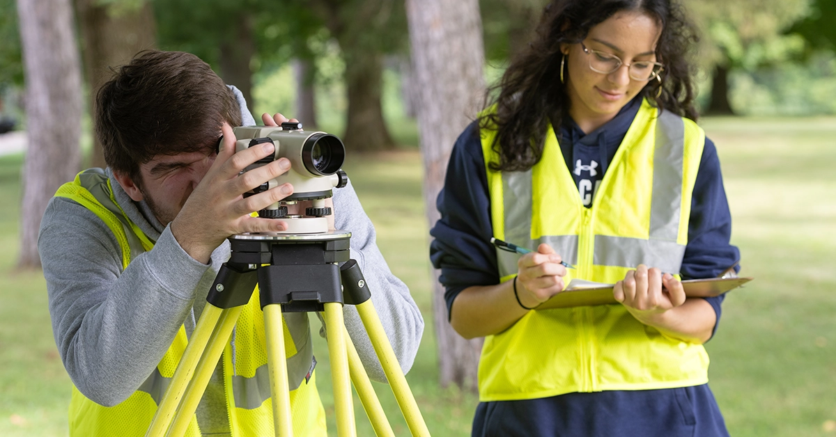 A student in a yellow vest looks through surveying scopes while another student also wearing a yellow vest takes notes.