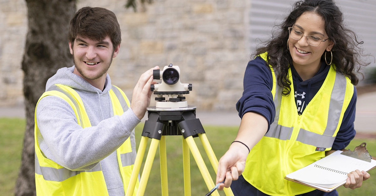 Two students with yellow safety vests prepare surveying equipment outside.