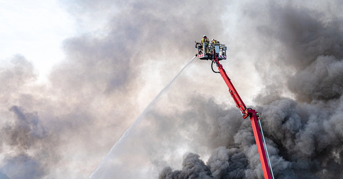 A firefighter sprays a large fire from a suspended ladder truck.