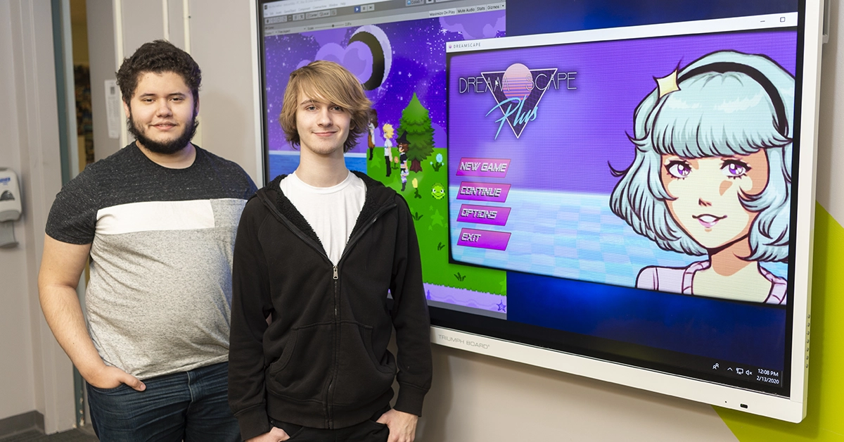 A large screen displays a video game rendering while two student creators stand in front.