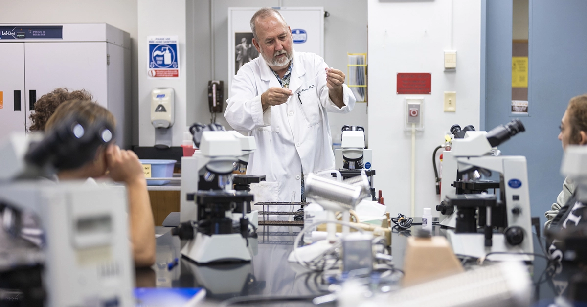 A professor demos a biology experiment while students prepare their microscopes in the bio lab.