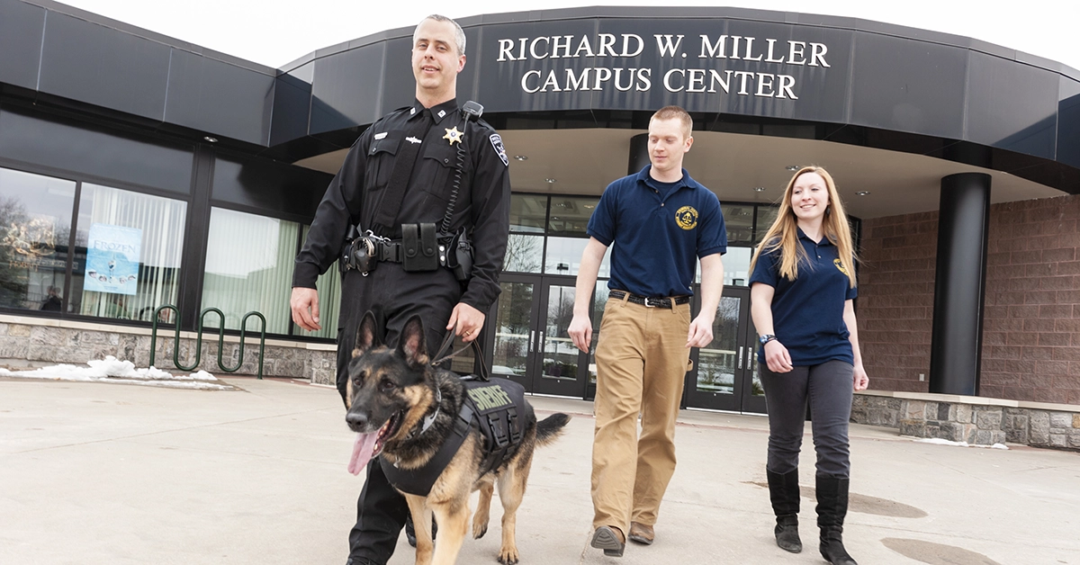 An officer walking a K-9 with two students outside the Miller Campus Center.