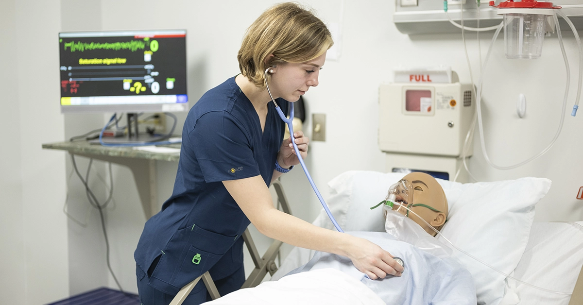 A nursing student checks the heart of a simulation dummy with a stethoscope in the Nursing lab.