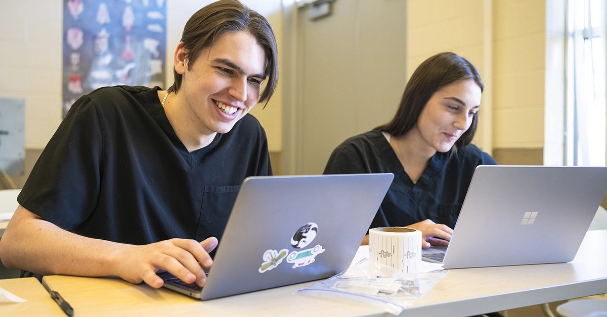 Two vet service students type on laptops in a classroom.