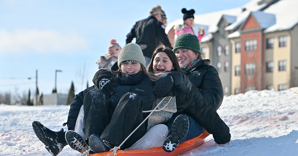 Three students sled down the hill near Kennedy Hall on a sunny winter day.