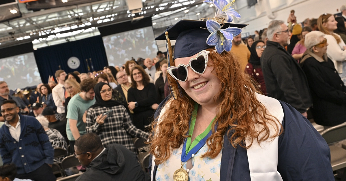 A graduate wearing sparkly heart sunglasses and adorned with purple lilies smiles from the audience.