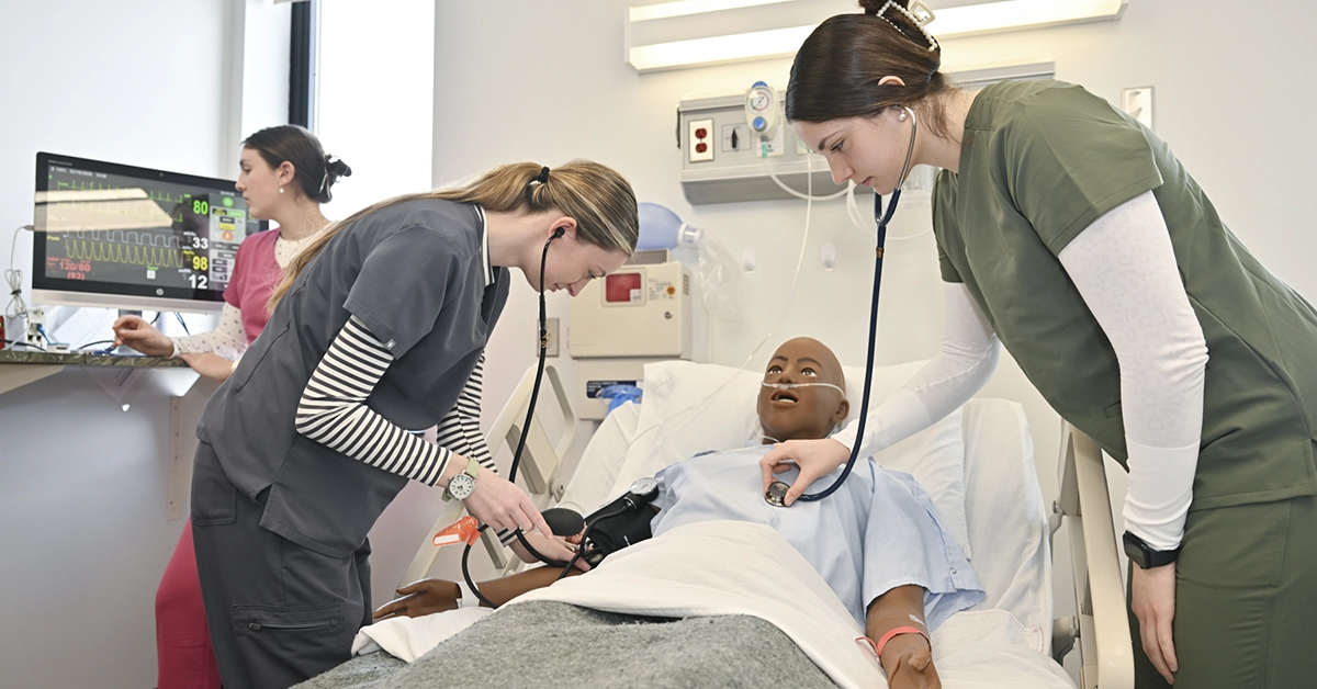 Three nursing students participate check vitals during a bedside simulation lab.