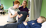 A student stretches a patient in the Physical Therapist lab while Instructional Support Associate Anne Reilly looks on.
