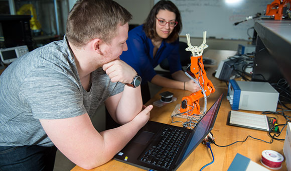 Two students program an orange robotic arm.