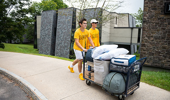 Two students with yellow shirts transport belongings on a cart.