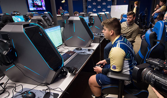 A students plays FIFA 19 while teammates and reporters look on.