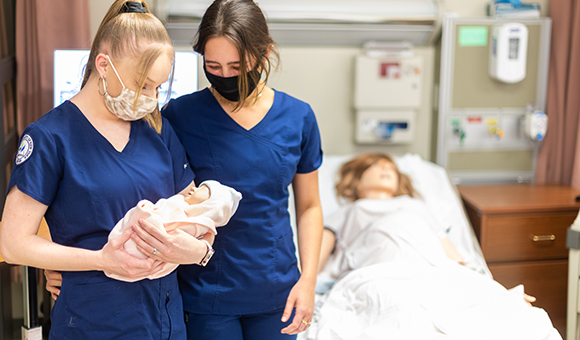 Nursing Students Shannon Szafranski and Thea Krywanczyk holding a swaddled baby manikin during a birthing simulation lab.