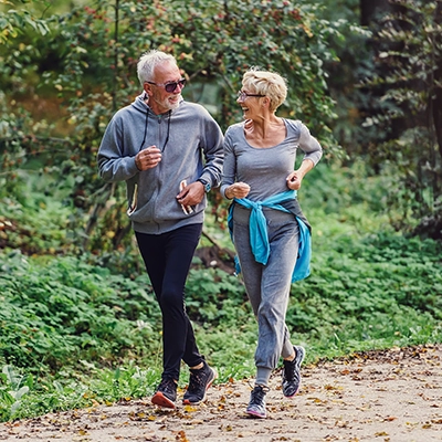 Two older adults jog together on a wooded trail.