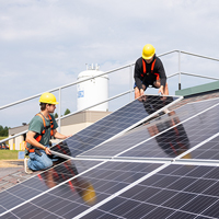 Two students wearing yellow hard hats install a solar panel on a mock roof.