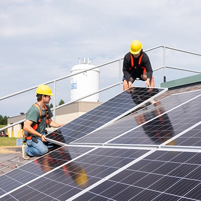 Two students wearing yellow hard hats install a solar panel on a mock roof.